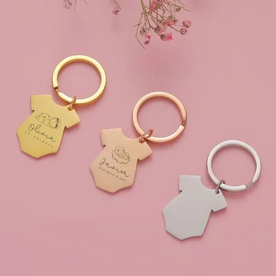 Llaveros forma de body de bebé, personalizados, en tonos metálicos sobre fondo rosa.