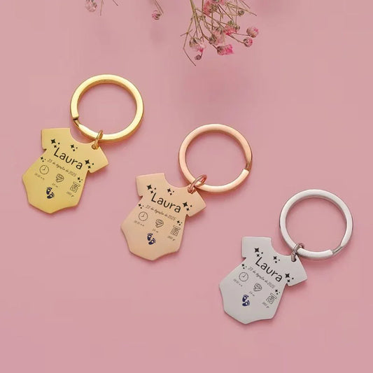 Llavero personalizado en forma de body para bebé en dorado, rosa y plateado sobre fondo rosa.