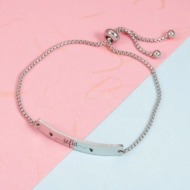 Pulsera personalizada de plata con nombre y corazones sobre fondo rosa y azul, ideal regalo divertido.